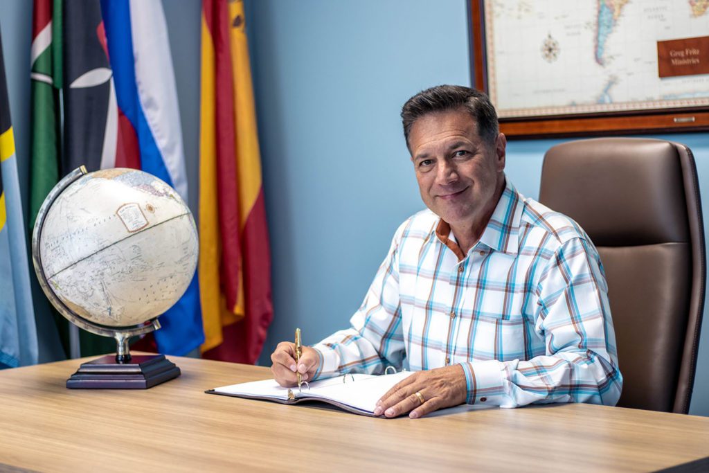 Greg Fritz sitting at a desk next to a globe, with the flags of different countries in the background